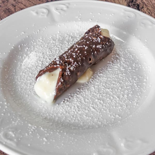 A chocolate-dipped cannoli filled with cream, dusted with powdered sugar on a white plate.