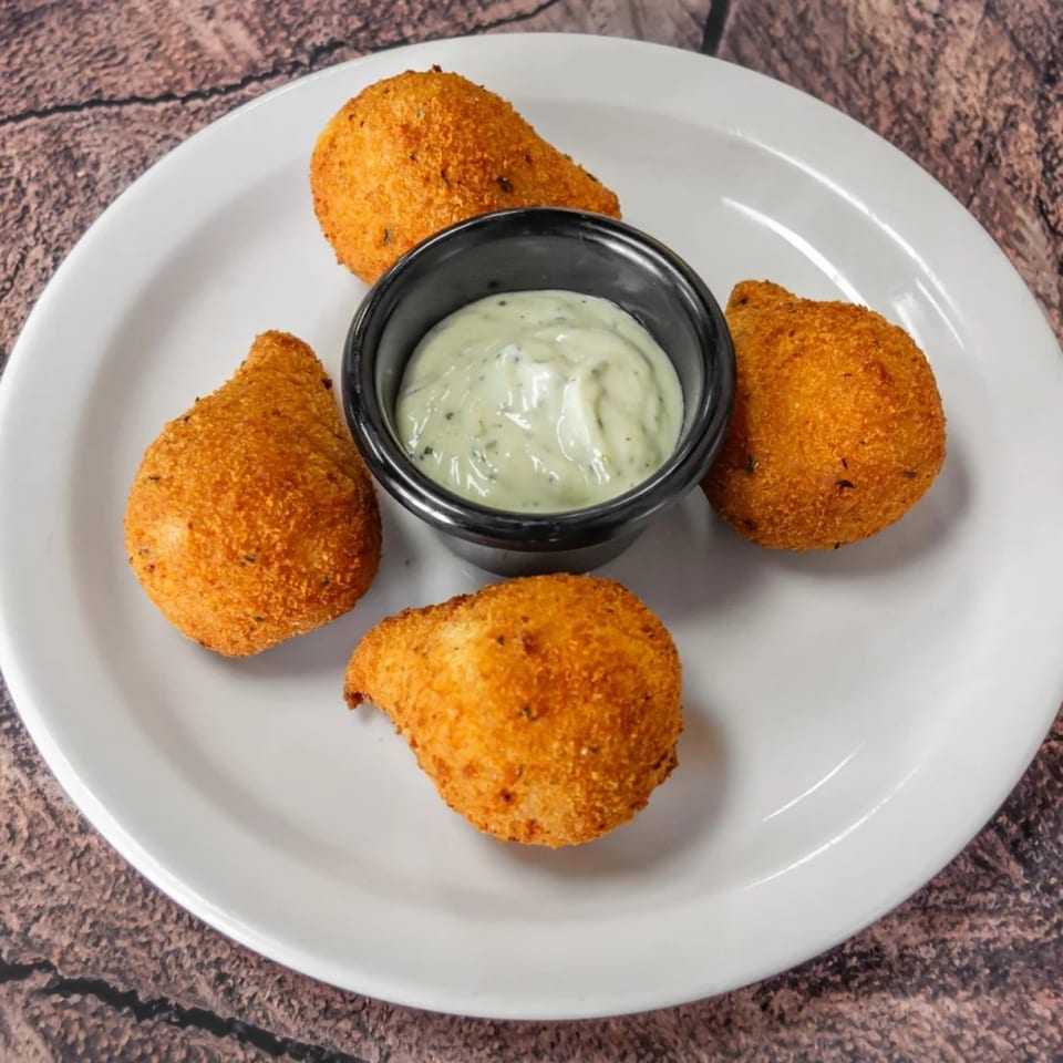Four golden-brown coxinha appetizers with a side of creamy green dipping sauce on a white plate.