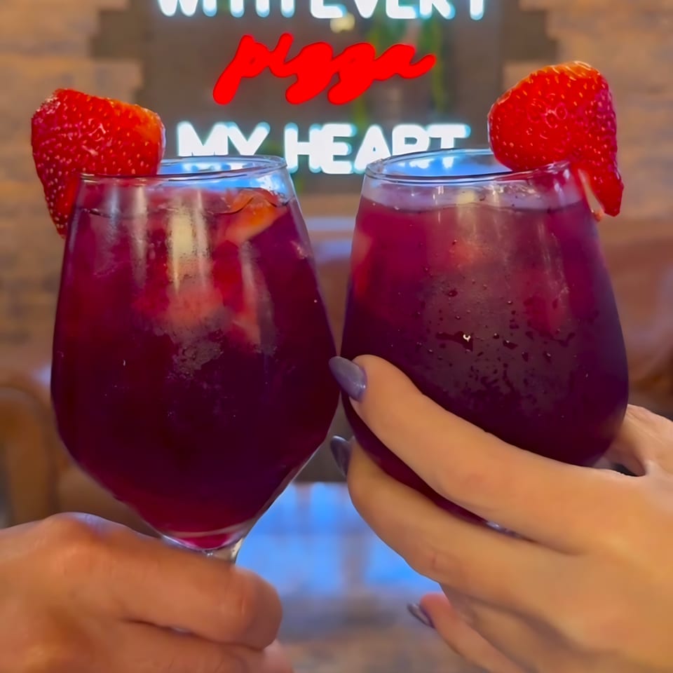 Two hands holding glasses of red sangria garnished with strawberries, with a neon sign in the background.