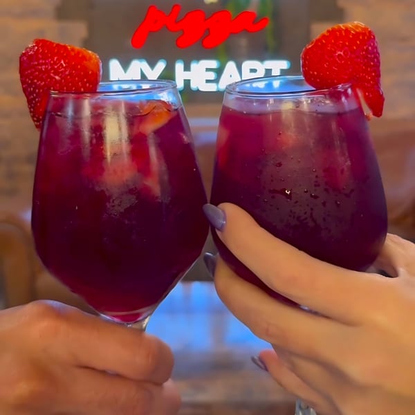 Two hands holding glasses of red sangria garnished with strawberries, toasting in front of a neon sign.